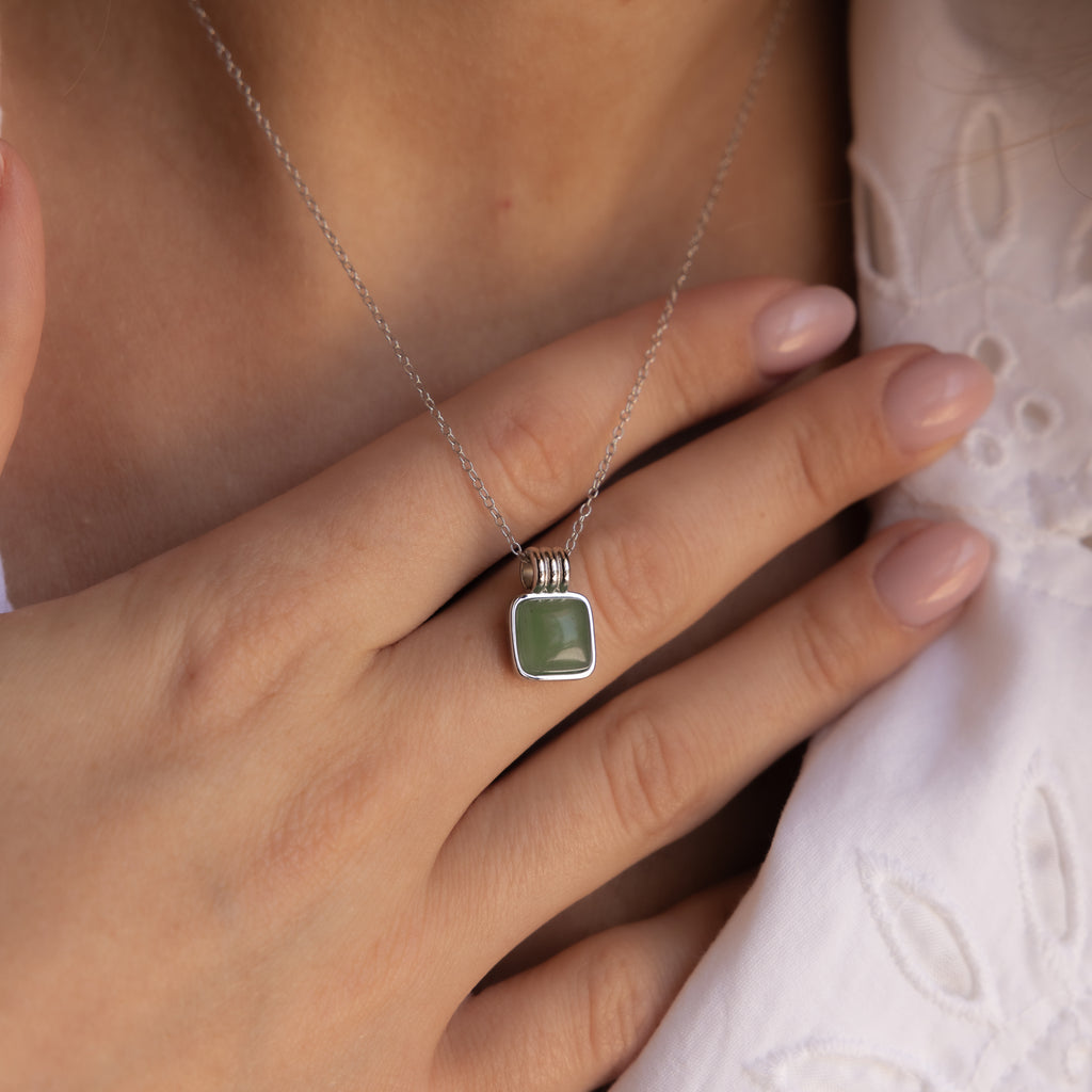 A person wearing a Jade Pendant Necklace in Silver, featuring a shimmering green jade stone, holds it near their collarbone for luck.