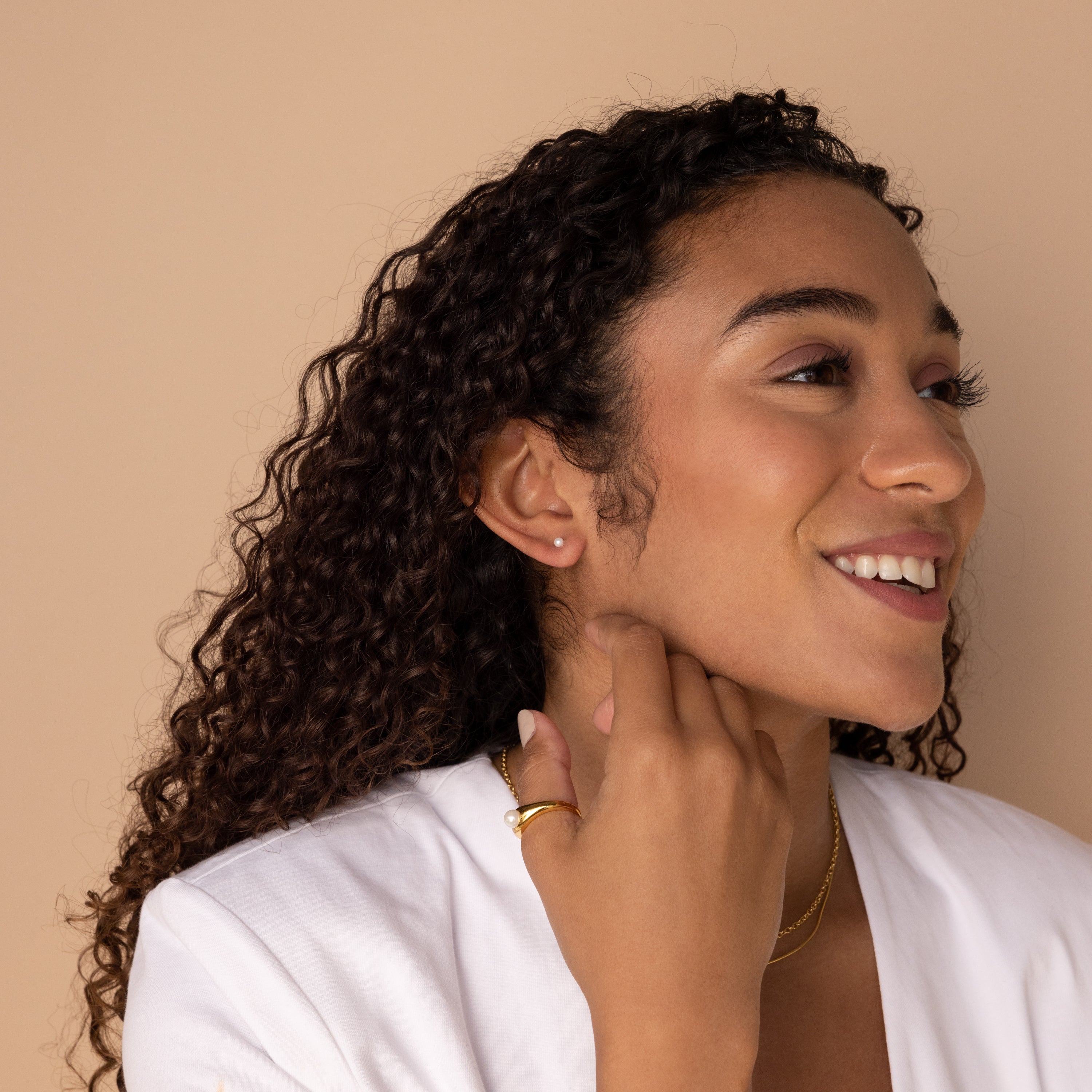 A smiling woman with curly hair wears a white top and the Dainty Pearl Studs, complemented by gold jewelry against a beige background.