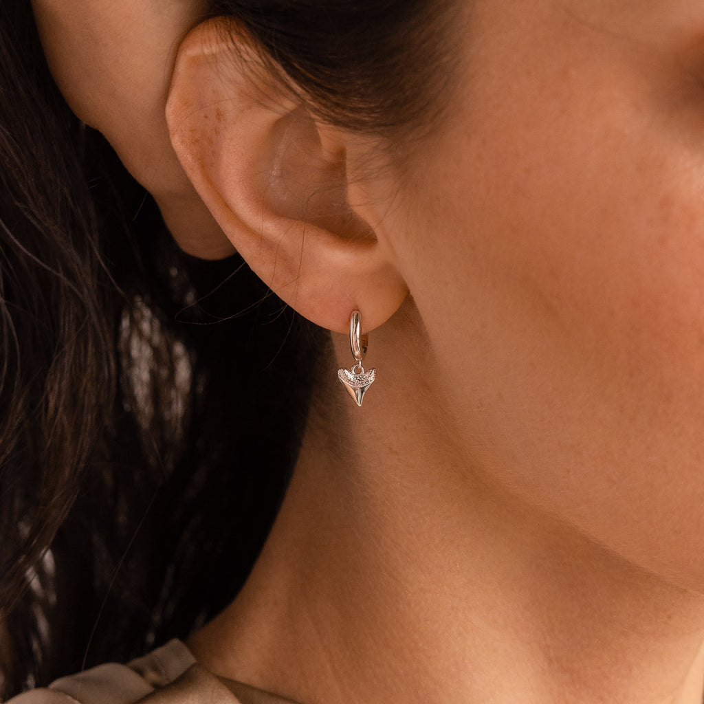 Close-up of a womans ear wearing Shark Tooth Huggies—small silver hoop earrings with a V-shaped pendant, perfect for adding subtle style to your next beach vacation.