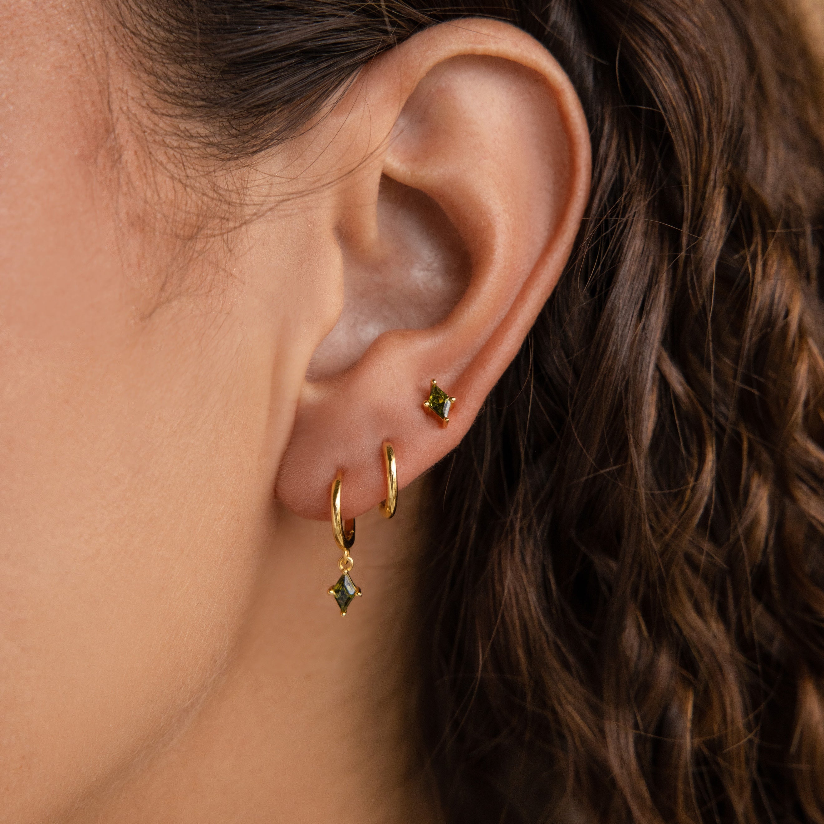 Close-up of an ear wearing the Peridot Earrings Set in 18K Gold—two gold hoops and a gold stud, each with green gemstones—ideal for earring stacking or as a chic cartilage earring.