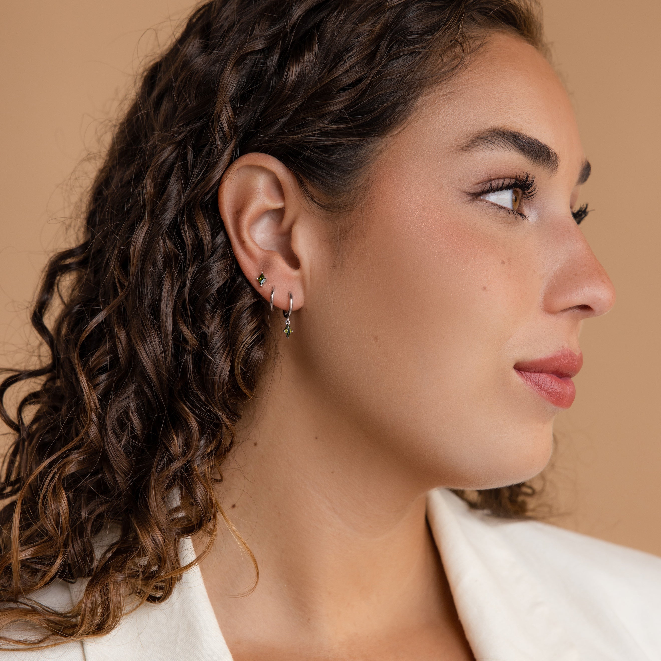 Woman with curly brown hair wears the Peridot Earrings Set in Sterling Silver, looking to the side against a beige background.