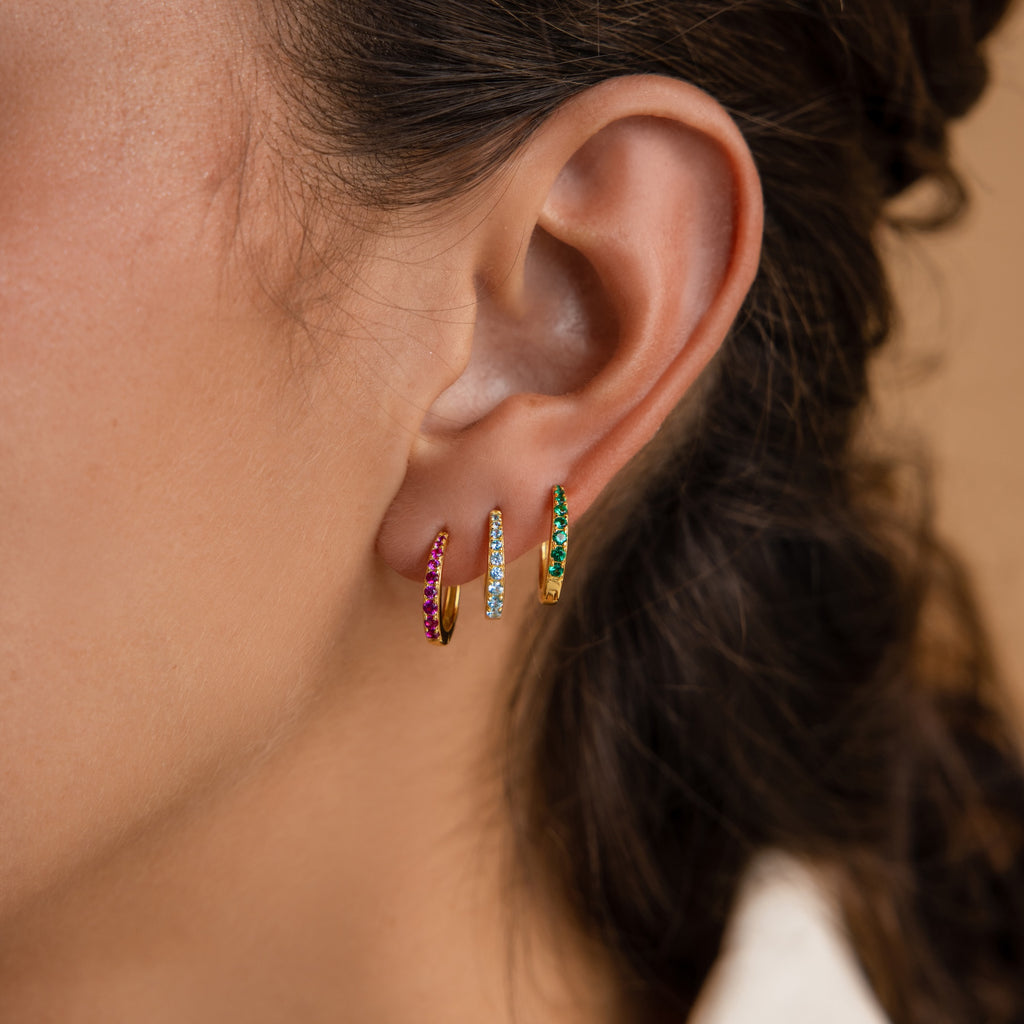 Close-up of an ear adorned with three Vera Pave Birthstone Huggies, each gold hoop featuring red, blue, and green gemstones for a vibrant look.