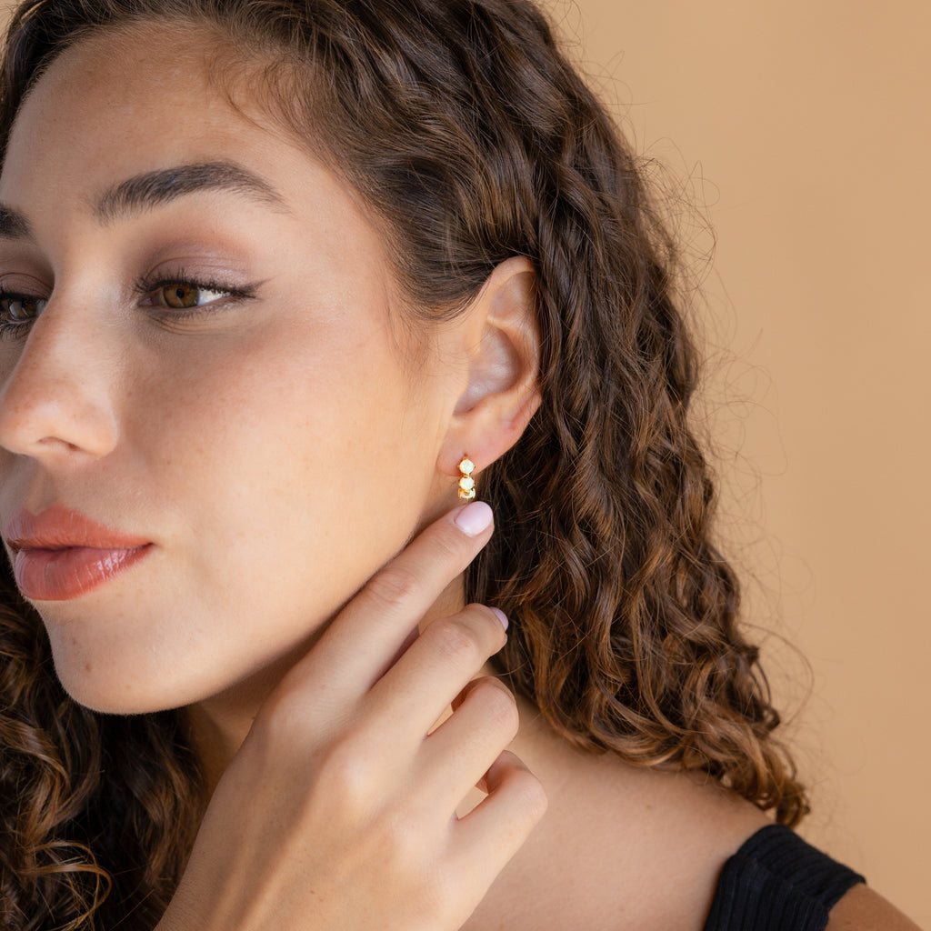 Woman with curly hair touches her Triple Aquamarine Huggies, minimalist hoop earrings, while gazing to the side against a beige background.