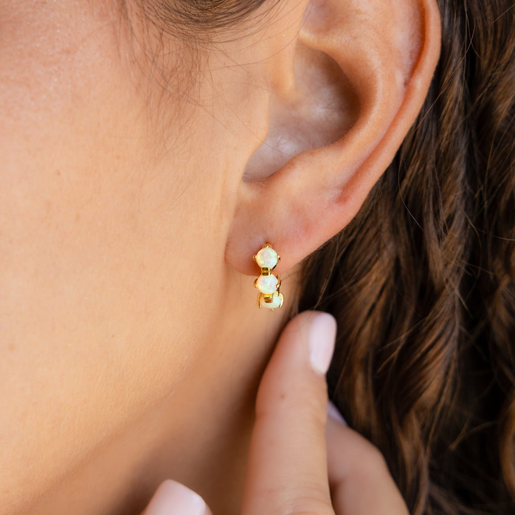 Close-up of a woman's ear wearing gold Triple Blue Zircon Huggies with three blue zircons, hand near her ear—perfect customizable gemstone earrings for a minimalist look.