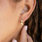 Close-up of an ear wearing gold Triple Peridot Huggies adorned with three light green gemstones; a hand gently touches the earlobe.