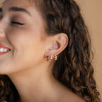 Smiling woman with curly hair wears colorful Triple Sapphire Huggies on a neutral background, highlighting personalized jewelry style.