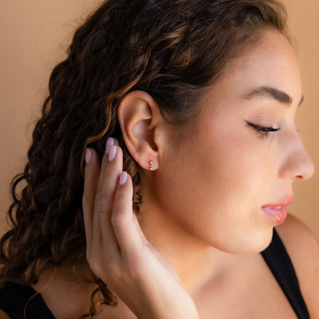A woman with curly hair touches her ear, showcasing petite gems from her Aquamarine Drop Studs against a beige background.
