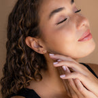 Woman with curly hair and closed eyes gently touching her face, wearing elegant Topaz Drop Studs and a black top—an exquisite accessory that adds a touch of sparkle.