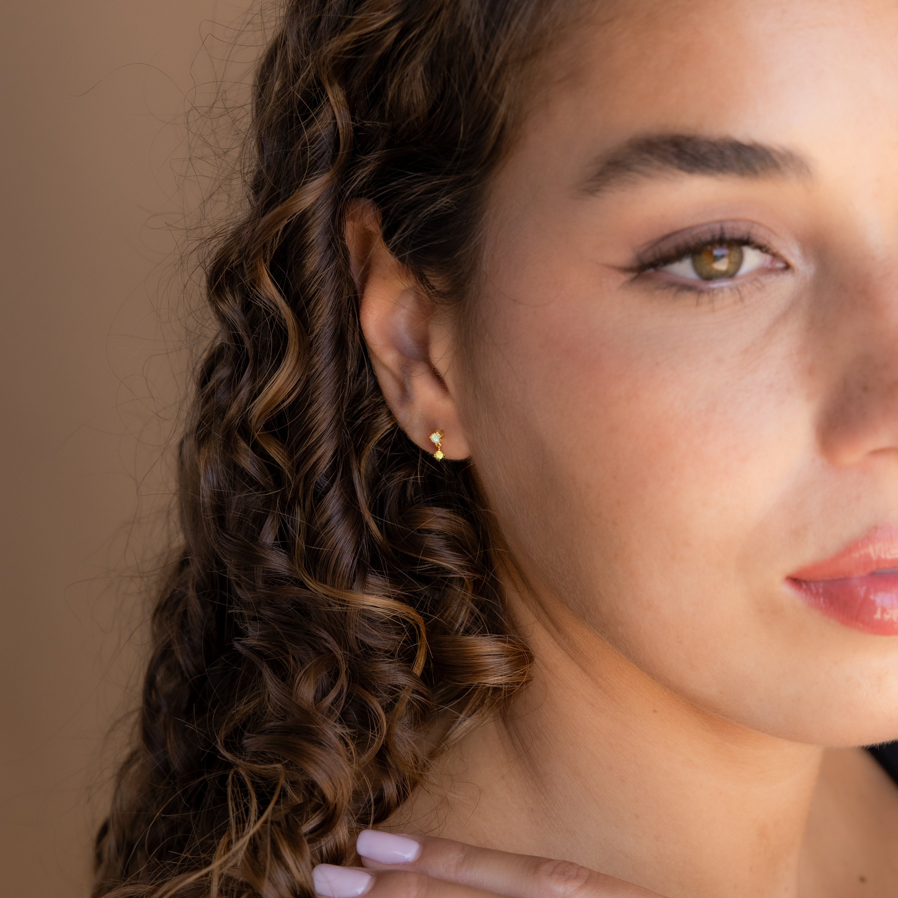 Woman with curly hair wearing Birthstone Drop Studs and a small gold hoop earring, gently touching her neck and gazing ahead.