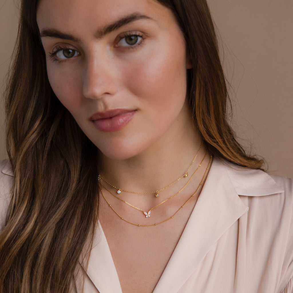 Woman with long brown hair wears a Pink Opal Butterfly Necklace in 18K Gold, layered with gold jewelry and a light pink blouse, looking at the camera.