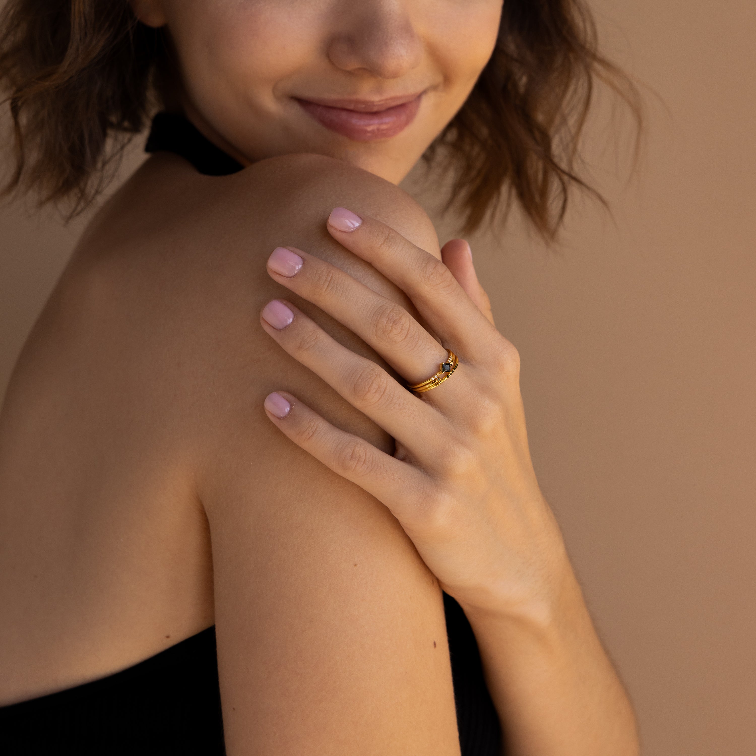 A woman smiles with her hand on her shoulder, wearing the Black Abyss Ring Set—a gold dainty band adorned with black gemstones—paired with light pink nail polish.