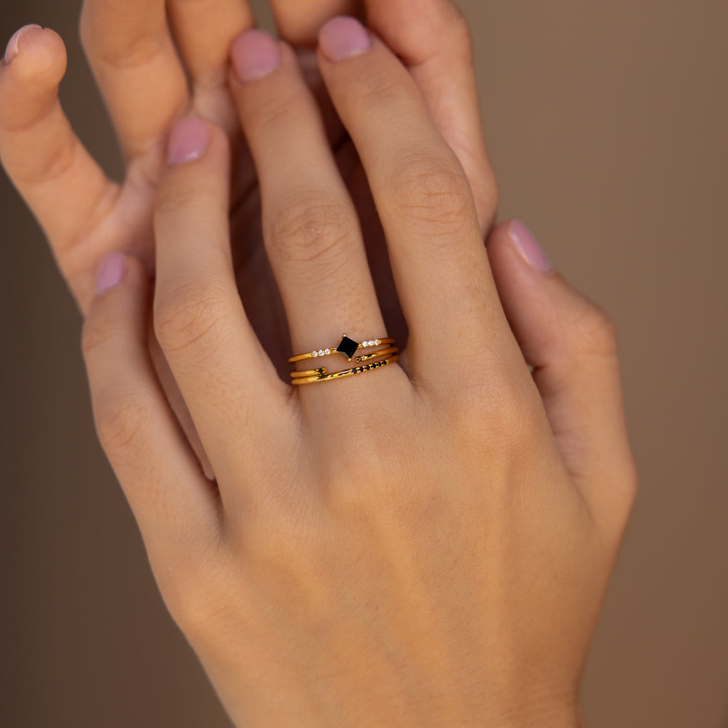 A hand with light pink nails wears the Black Abyss Ring Set—three stacked gold rings with dainty bands and a small black gemstone.