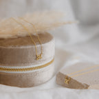 The Mellow Dainty Initial Necklace with a gold letter K pendant rests on a beige velvet jewelry box, next to another custom accessory displayed on a stone surface.