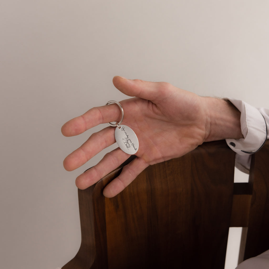 A hand holds the Handwriting Oval Keychain, featuring a small personalized tag, resting on the back of a wooden chair.