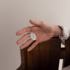 A hand holds the Handwriting Oval Keychain, featuring a small personalized tag, resting on the back of a wooden chair.