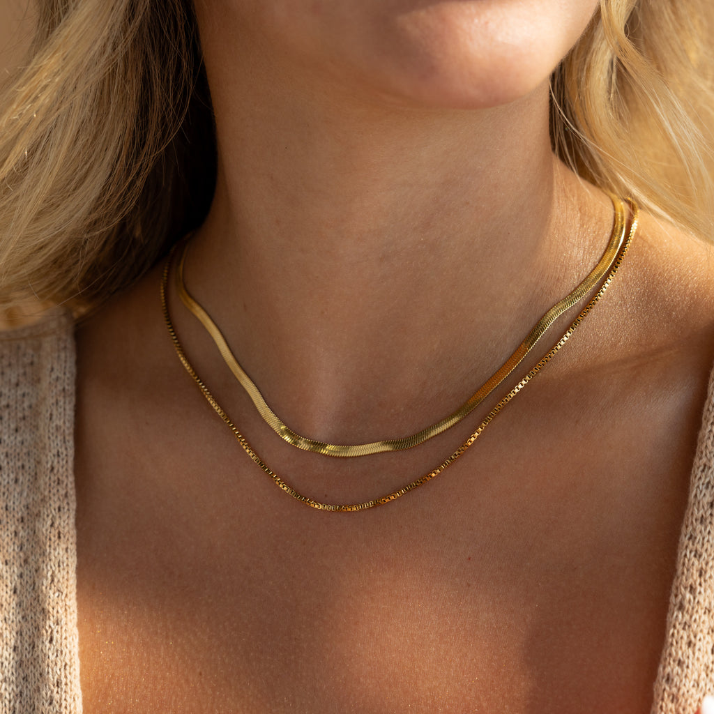 A woman wears the Herringbone Duo Chain Necklace, featuring layered gold herringbone and beaded chains, styled over a light top.