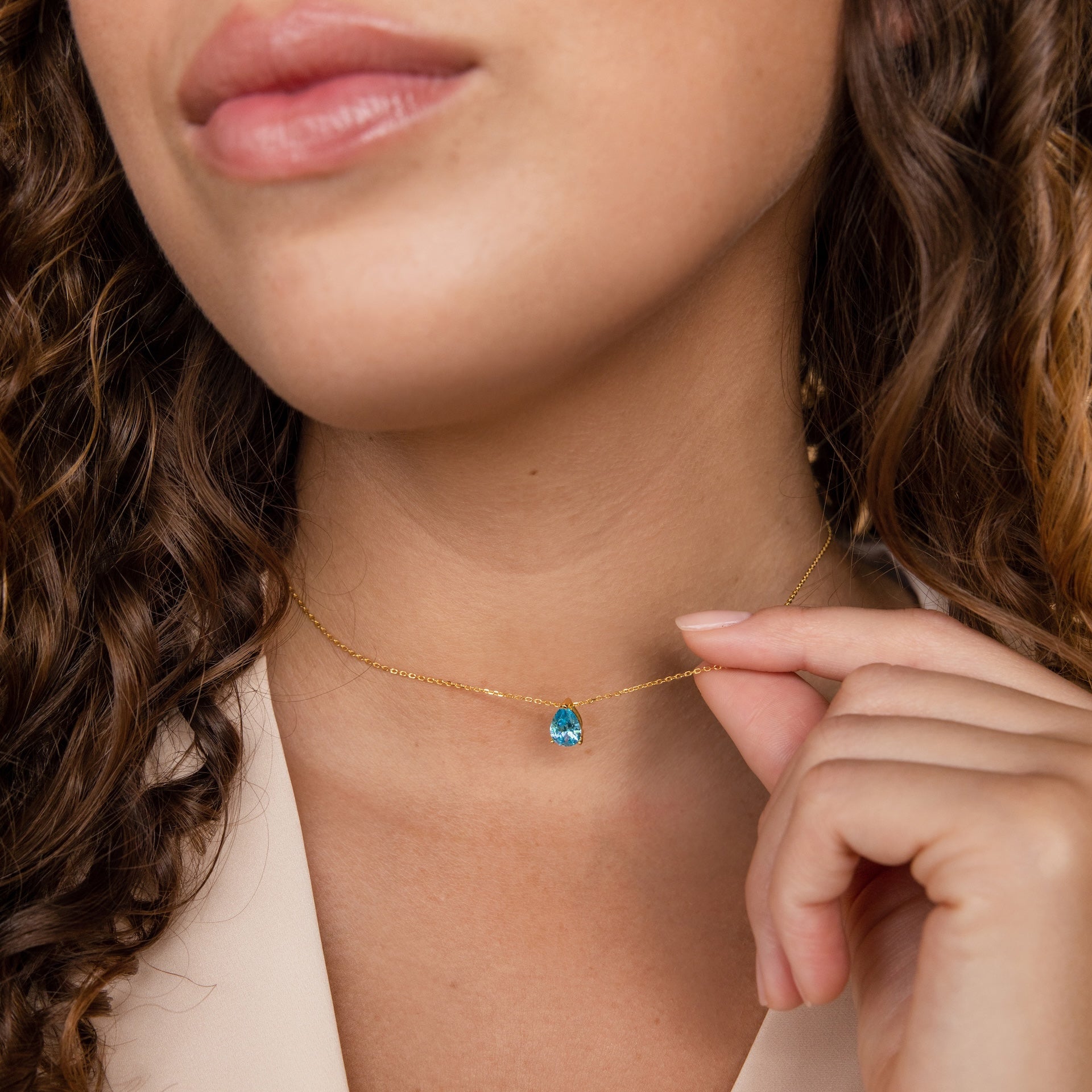 A woman with curly hair gently holds the Ophelia Blue Zircon Necklace, featuring a gold chain and blue gemstone pendant, near her neck.