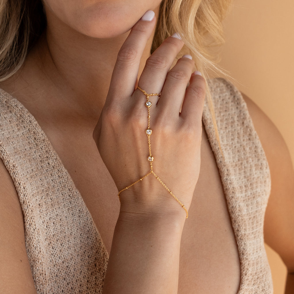 A woman wears the Diamond Bezel Hand Chain, adorned with small gemstone accents, as she rests her hand on her chin.