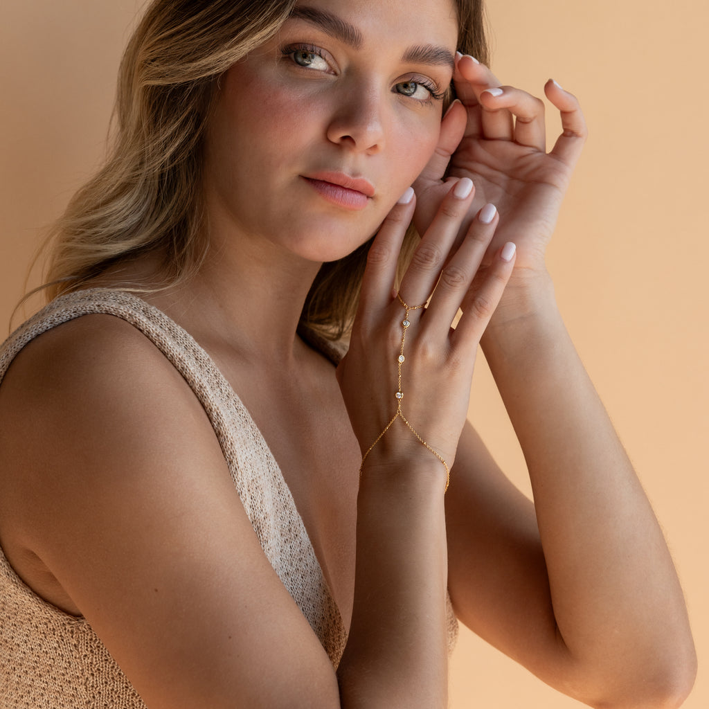 A woman wears the Diamond Bezel Hand Chain in 18K Gold, styled with a beige sleeveless top against a peach background.
