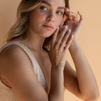 A woman wears the Diamond Bezel Hand Chain in 18K Gold, styled with a beige sleeveless top against a peach background.