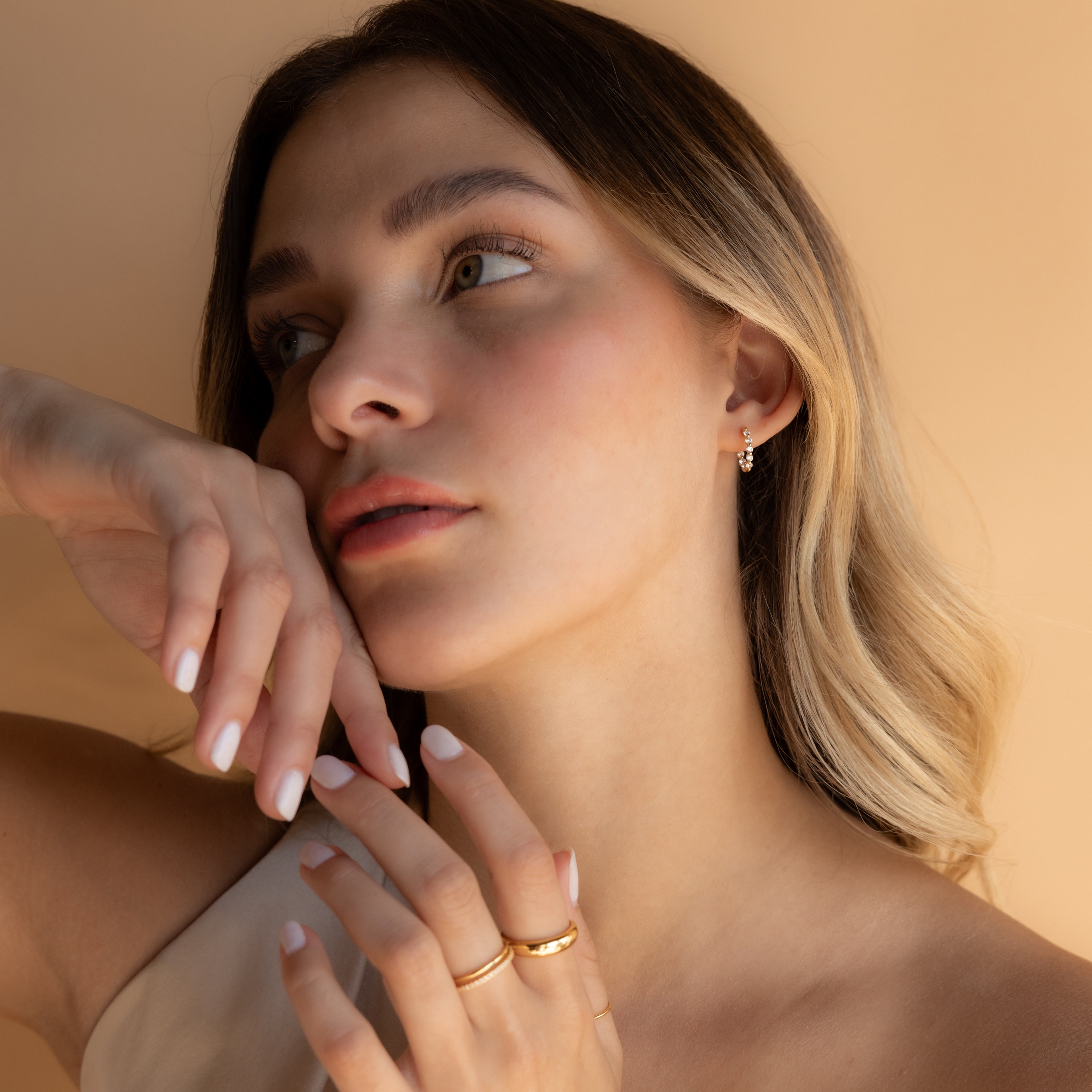 A woman with light brown hair poses thoughtfully against a beige background, wearing Inside Out Diamond Huggies and gold jewelry, complemented by neutral makeup.