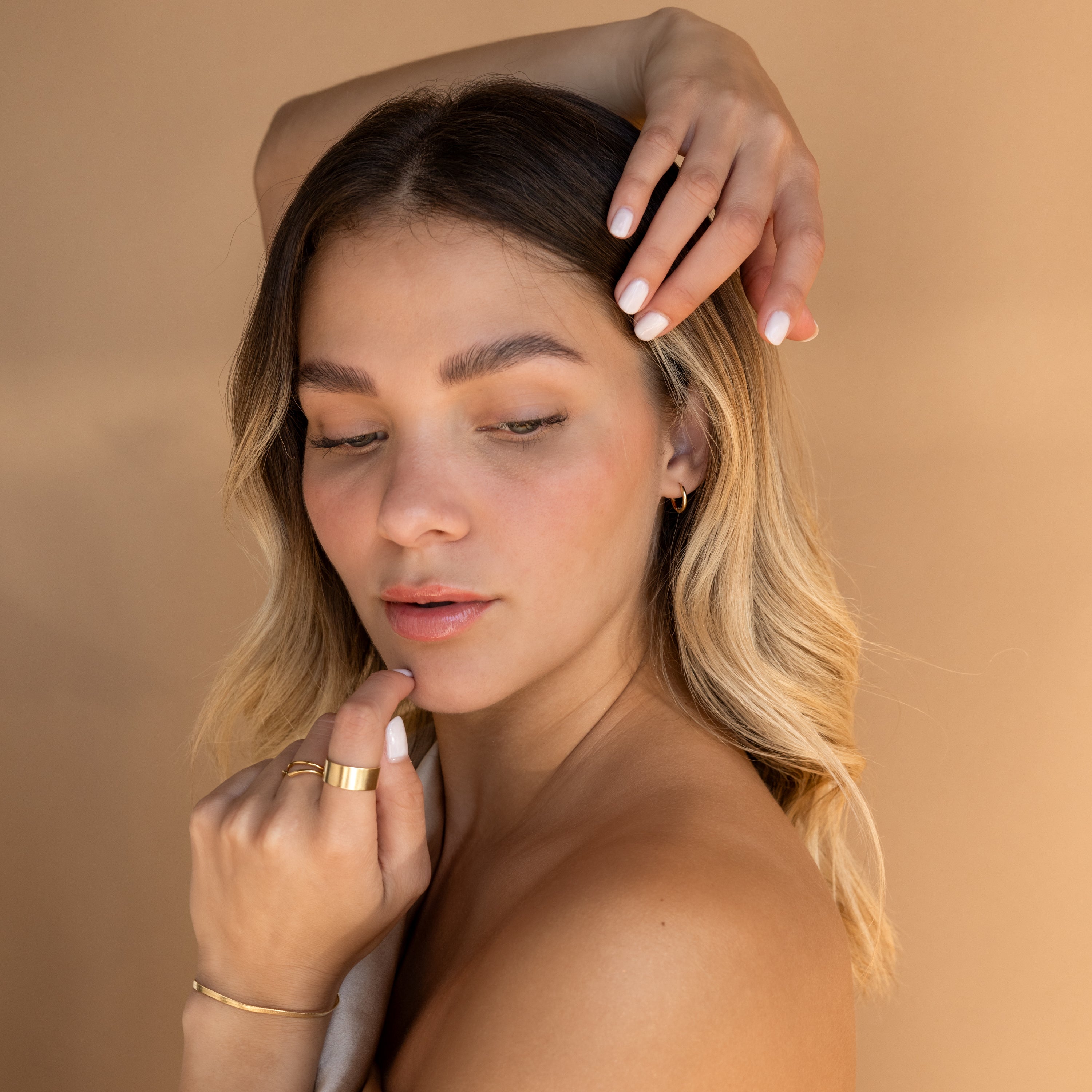 Woman with wavy blonde hair poses gracefully against a beige background, wearing Small Bold Hoops in Sterling Silver, gold rings, and natural makeup.