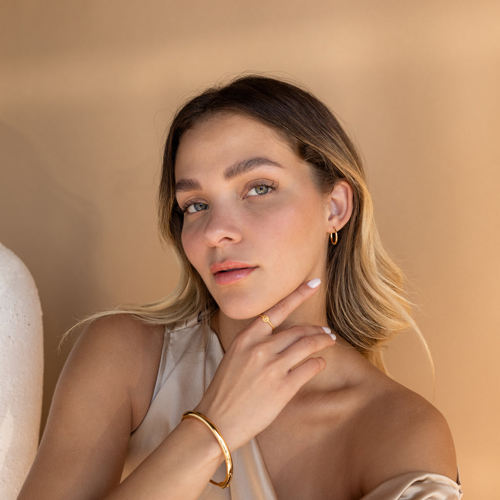 A woman with light hair poses against a beige background, her hand on her chin, effortlessly showcasing Medium Bold Hoops in Sterling Silver for an elegant everyday look.