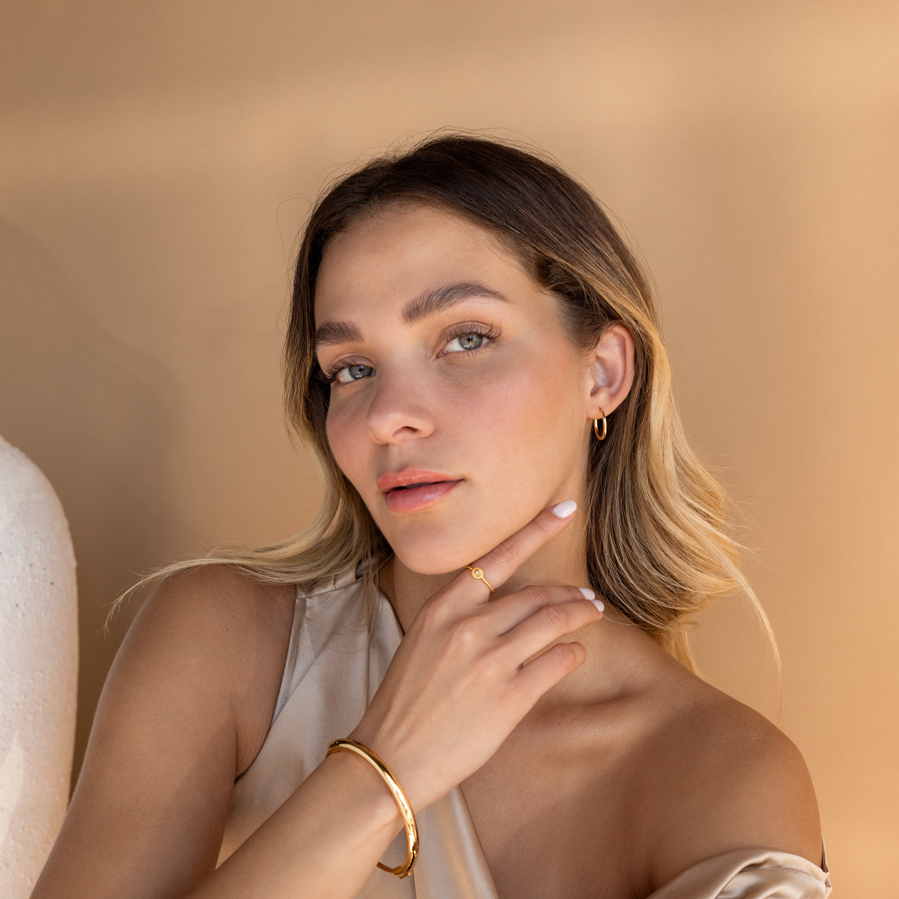 A woman with light hair poses against a beige background, her hand on her chin, effortlessly showcasing Medium Bold Hoops in Sterling Silver for an elegant everyday look.