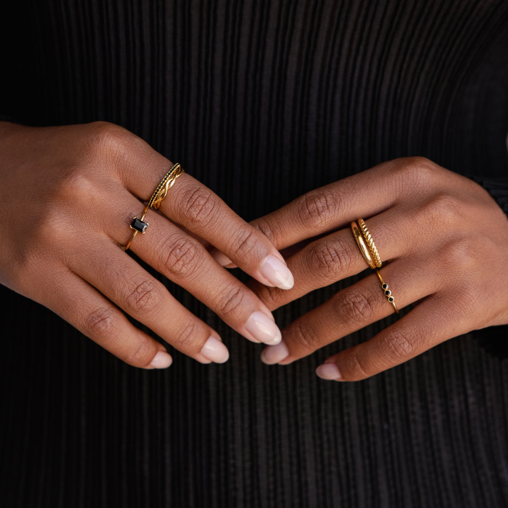 Two manicured hands wearing gold rings, including the Black Emerald Ring, are displayed against a black, textured background.