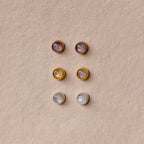 Six pairs of round stud earrings, including the Moonstone Bezel Studs, come in gold, silver, and rose gold settings for a subtle, ethereal look displayed on a beige background.