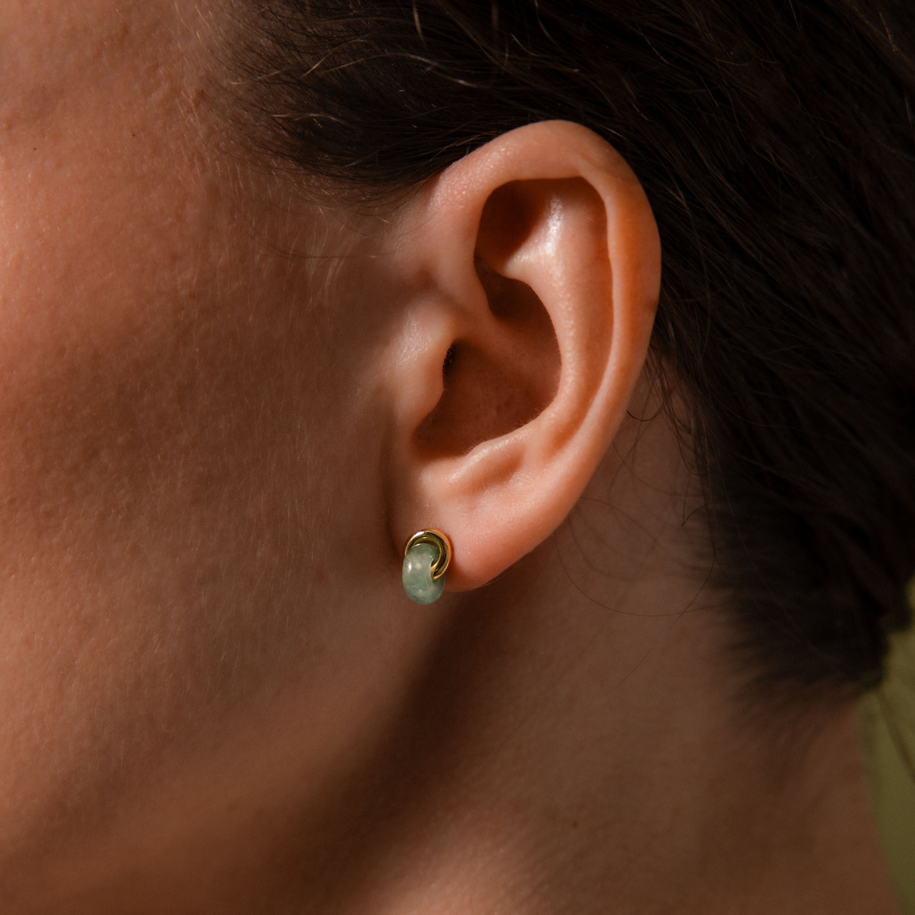 Close-up of a woman's ear wearing a small, smooth green jade stud earring with a gold halo setting.