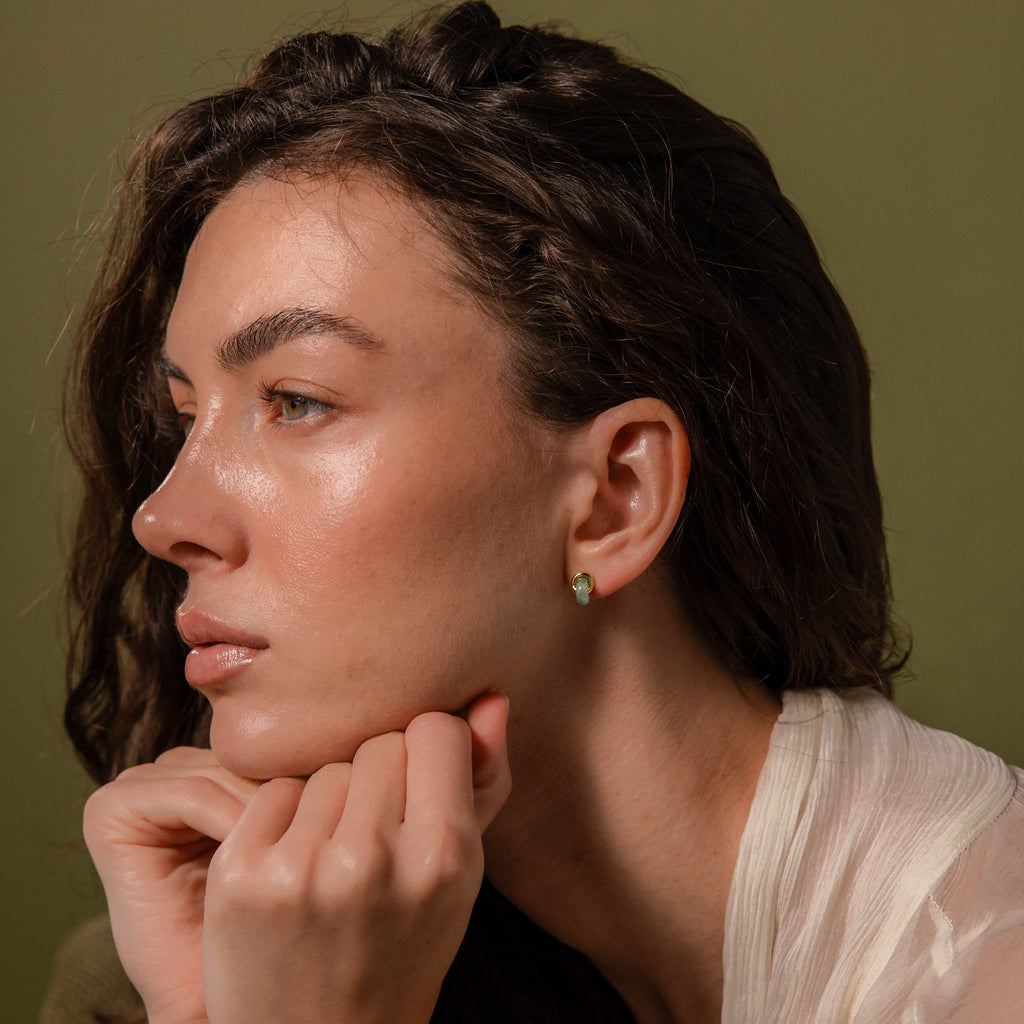 Woman wearing small, round green jade stud earrings with a gold halo, against a green background.