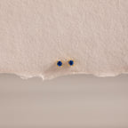 The Dainty Sapphire Flatback Studs feature small round blue gemstones, perfect for multiple piercings, and are displayed on textured beige paper.