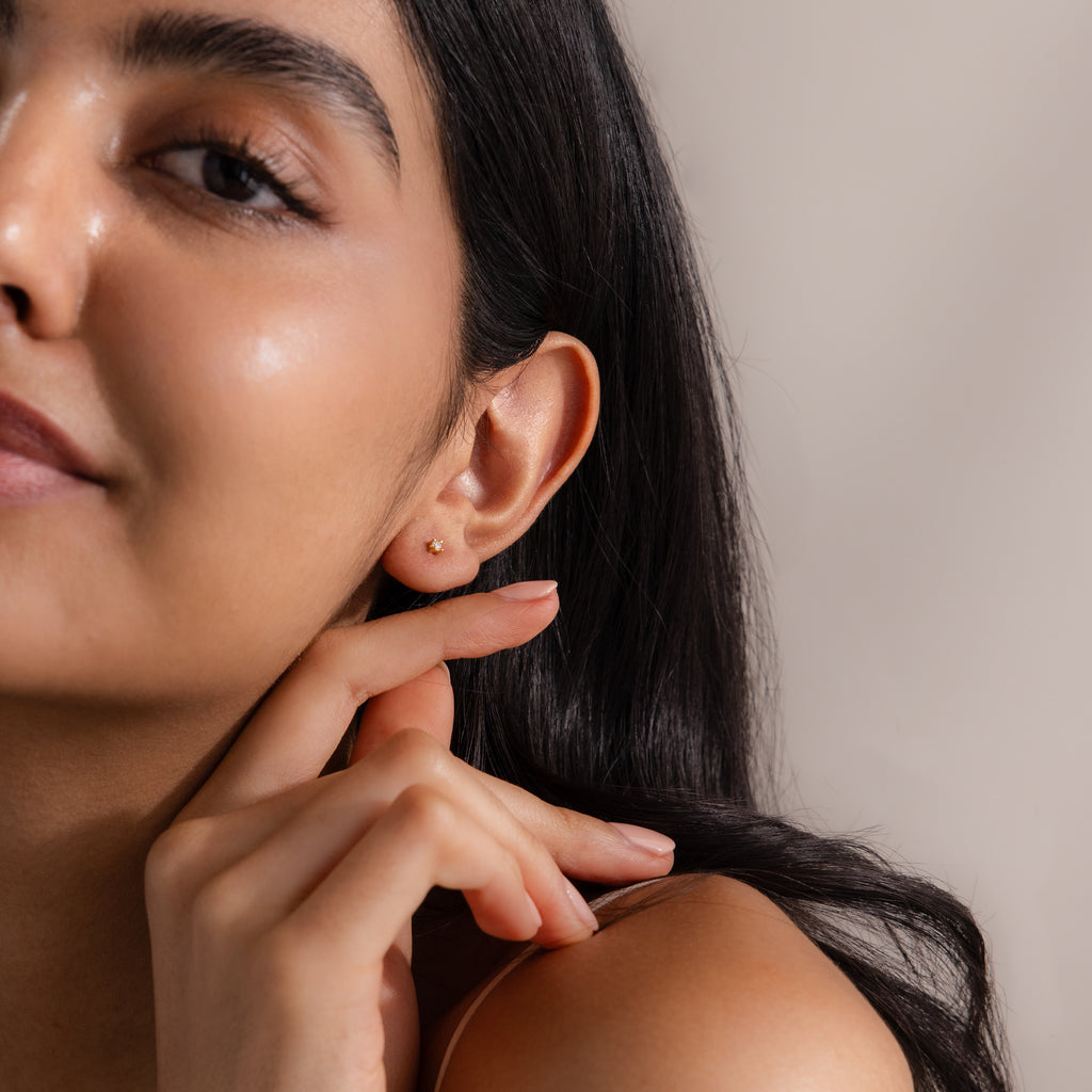 A woman with long dark hair touches her ear, wearing Dainty Birthstone Flatback Studs in soft natural lighting—ideal earrings for multiple piercings.