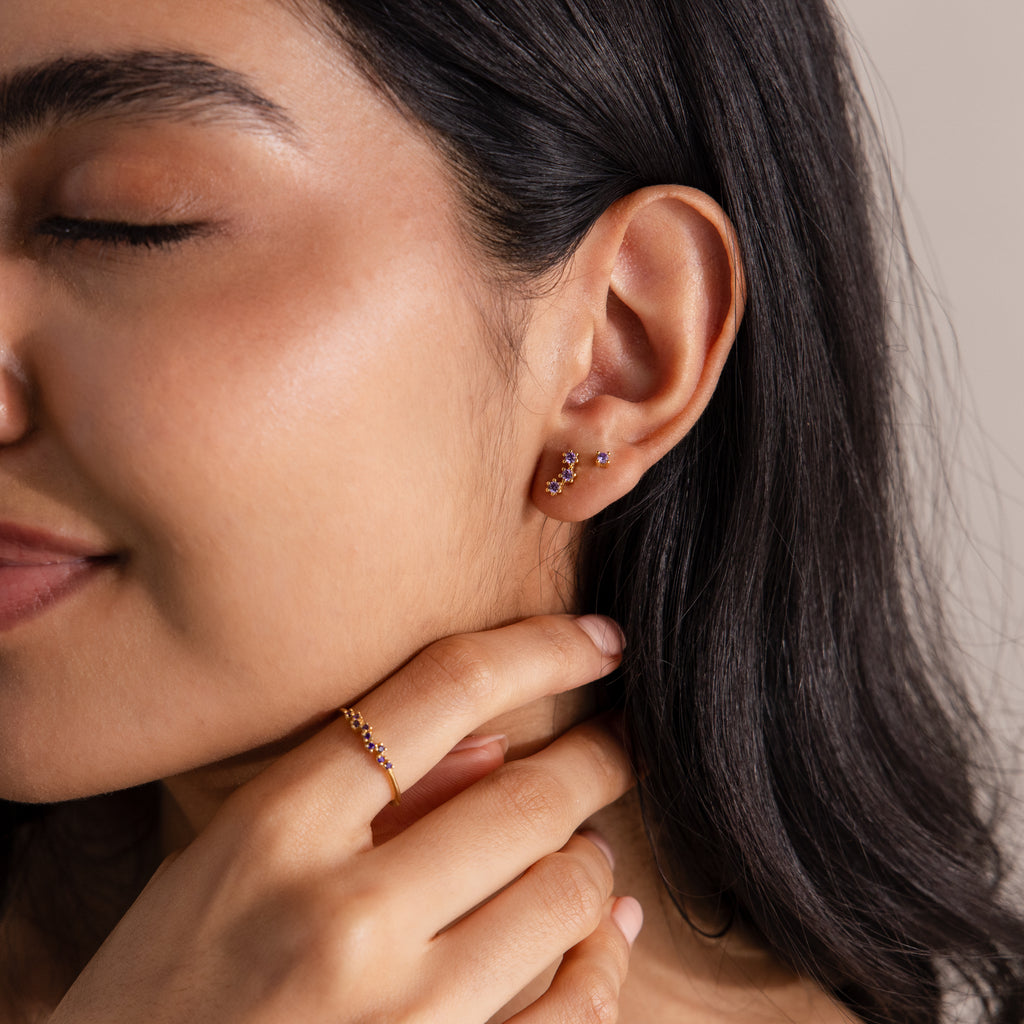 A woman with closed eyes gently touches her neck, wearing gold earrings, a matching gold ring, and Dainty Amethyst Flatback Studs for multiple piercings, giving her elegant style a modern twist.
