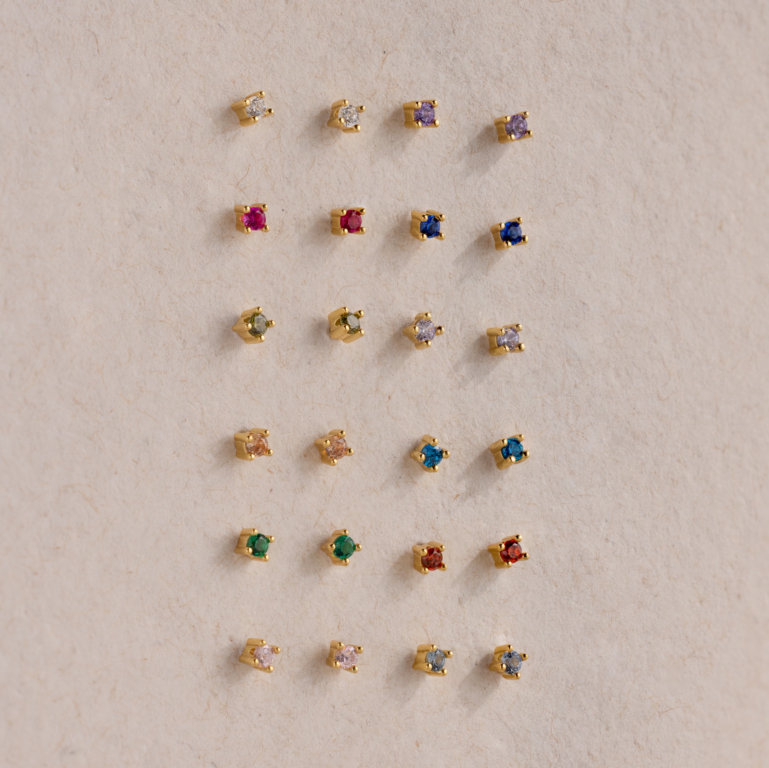 Twenty pairs of Dainty Amethyst Flatback Studs, ideal for multiple piercings, are displayed in a neat grid on a beige surface.
