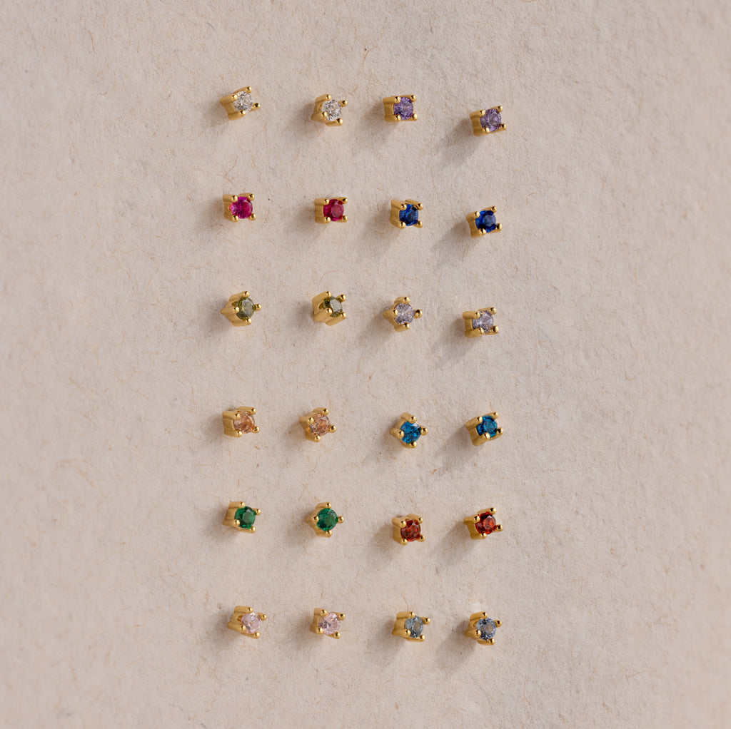 Nine pairs of Dainty Birthstone Flatback Studs, each with a unique colored stone, are displayed in a grid on a beige surface, creating a timeless style ideal for multiple piercings.
