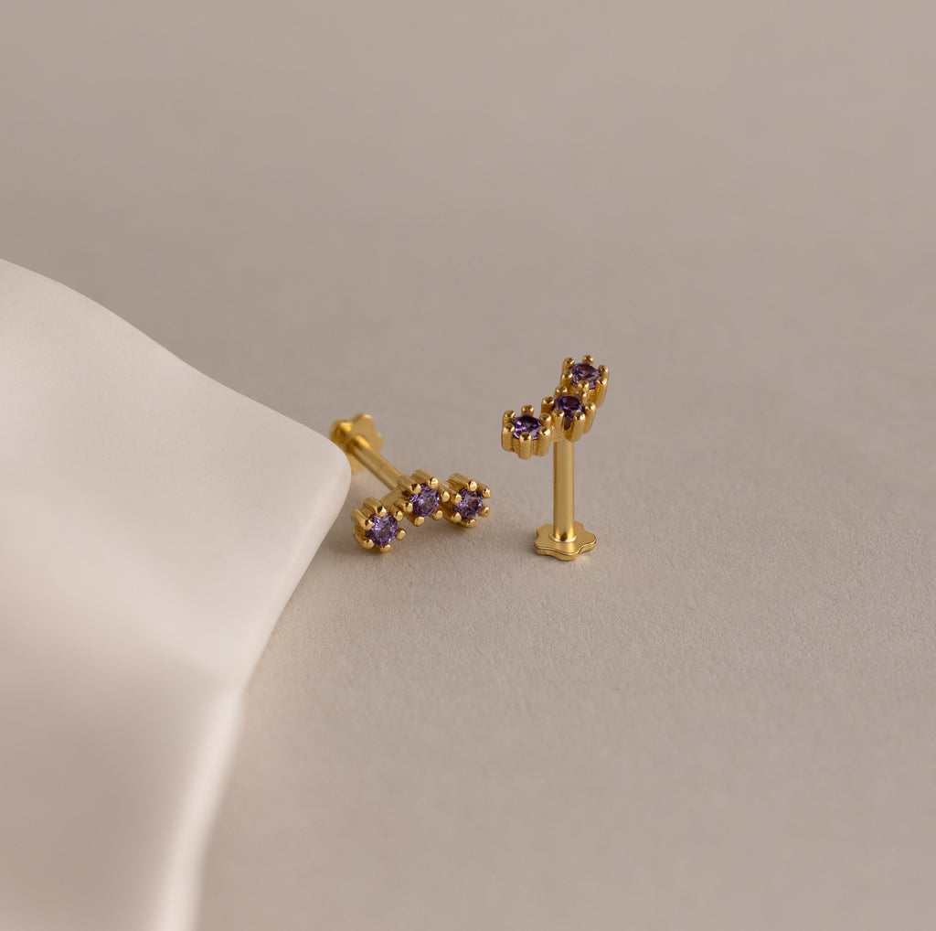 Alexandrite Cluster Flatback Studs featuring gold clusters and purple gemstones rest on a beige surface beside white fabric—ideal as personalized jewelry or birthstone earrings.