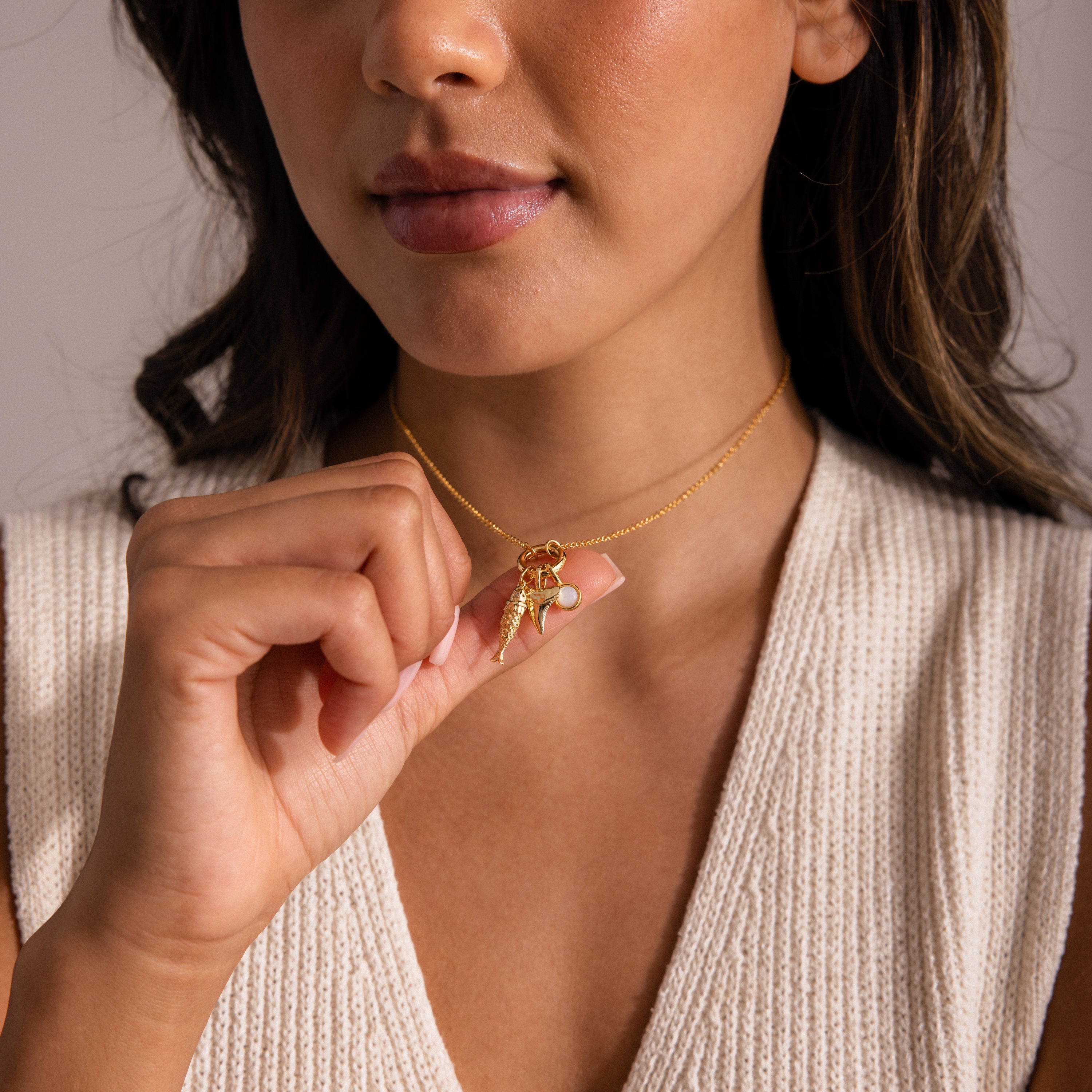Gold Fish Charm styled on a chain necklace with a pearl charm and seashell charm, held gently by model wearing a knit top.
