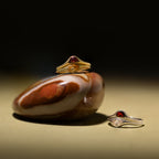 The Sculpted Toi et Moi Ring Set features two gold rings with red stones displayed on polished brown stone, alongside a sculpted silver ring adorned with a matching red stone.