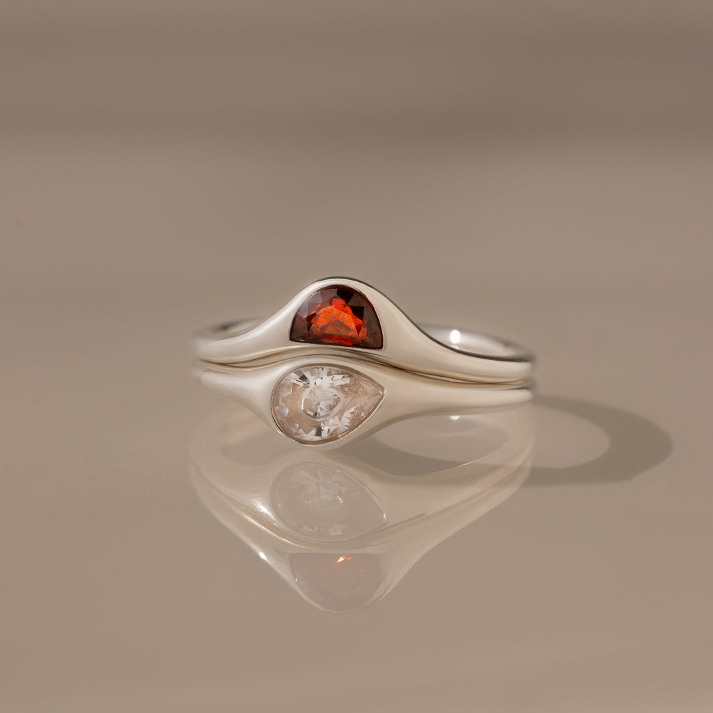The Sculpted Toi et Moi Ring Set in Silver features a red gemstone and a clear gemstone side by side, reflecting elegantly on a smooth surface.