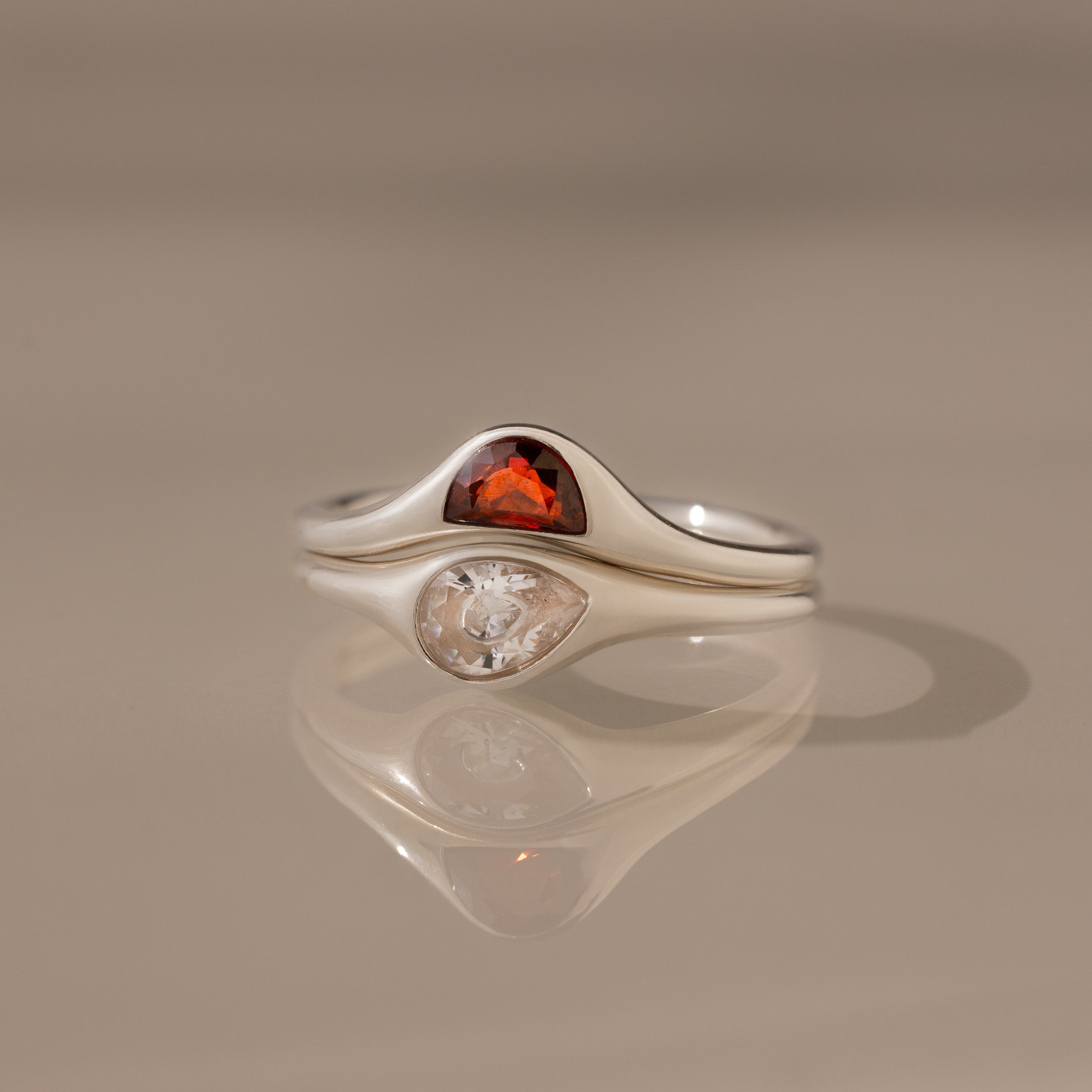 The Sculpted Toi et Moi Ring Set in Silver features a red gemstone and a clear gemstone side by side, reflecting elegantly on a smooth surface.