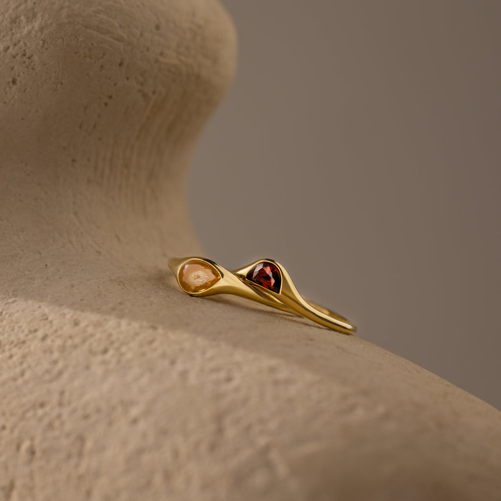 The Sculpted Toi et Moi Ring Set in 18K Gold, featuring two gemstones—one red and one peach—rests on a textured beige surface.