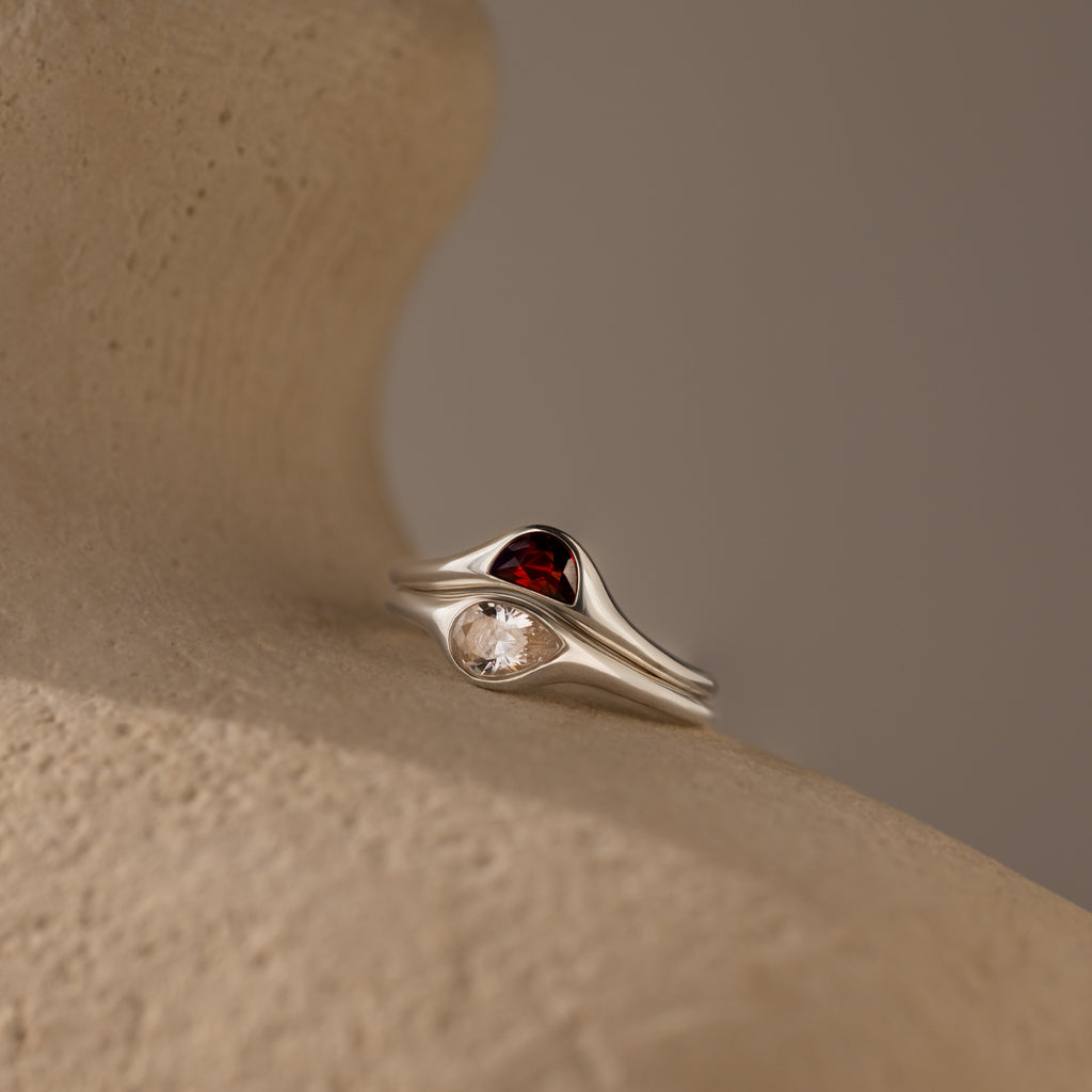 The Sculpted Toi et Moi Ring Set in Silver features red and clear gemstones, elegantly showcased on a textured beige surface under soft lighting.