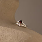 The Sculpted Toi et Moi Ring Set in Silver features red and clear gemstones, elegantly showcased on a textured beige surface under soft lighting.