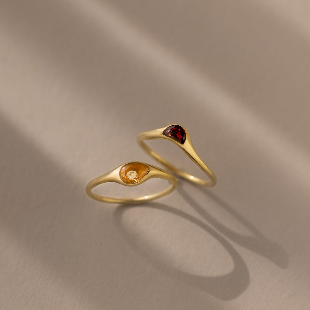 The Sculpted Toi et Moi Ring Set in 18K Gold features two gold rings with small teardrop-shaped amber and red gemstones, elegantly displayed on a beige surface.