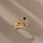 The Sculpted Toi et Moi Ring Set in 18K Gold features two gold rings with small teardrop-shaped amber and red gemstones, elegantly displayed on a beige surface.
