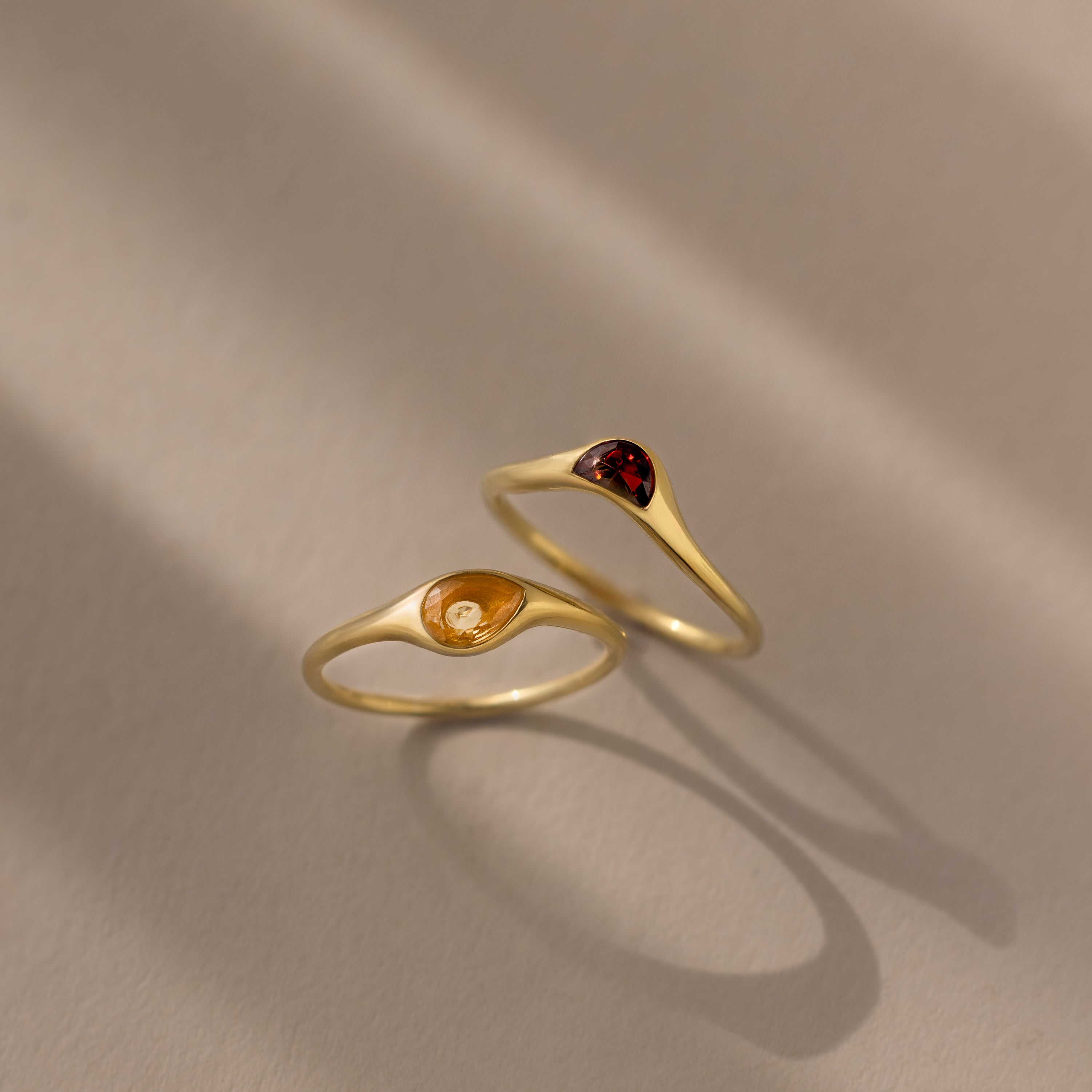 The Sculpted Toi et Moi Ring Set in 18K Gold features two gold rings with small teardrop-shaped amber and red gemstones, elegantly displayed on a beige surface.