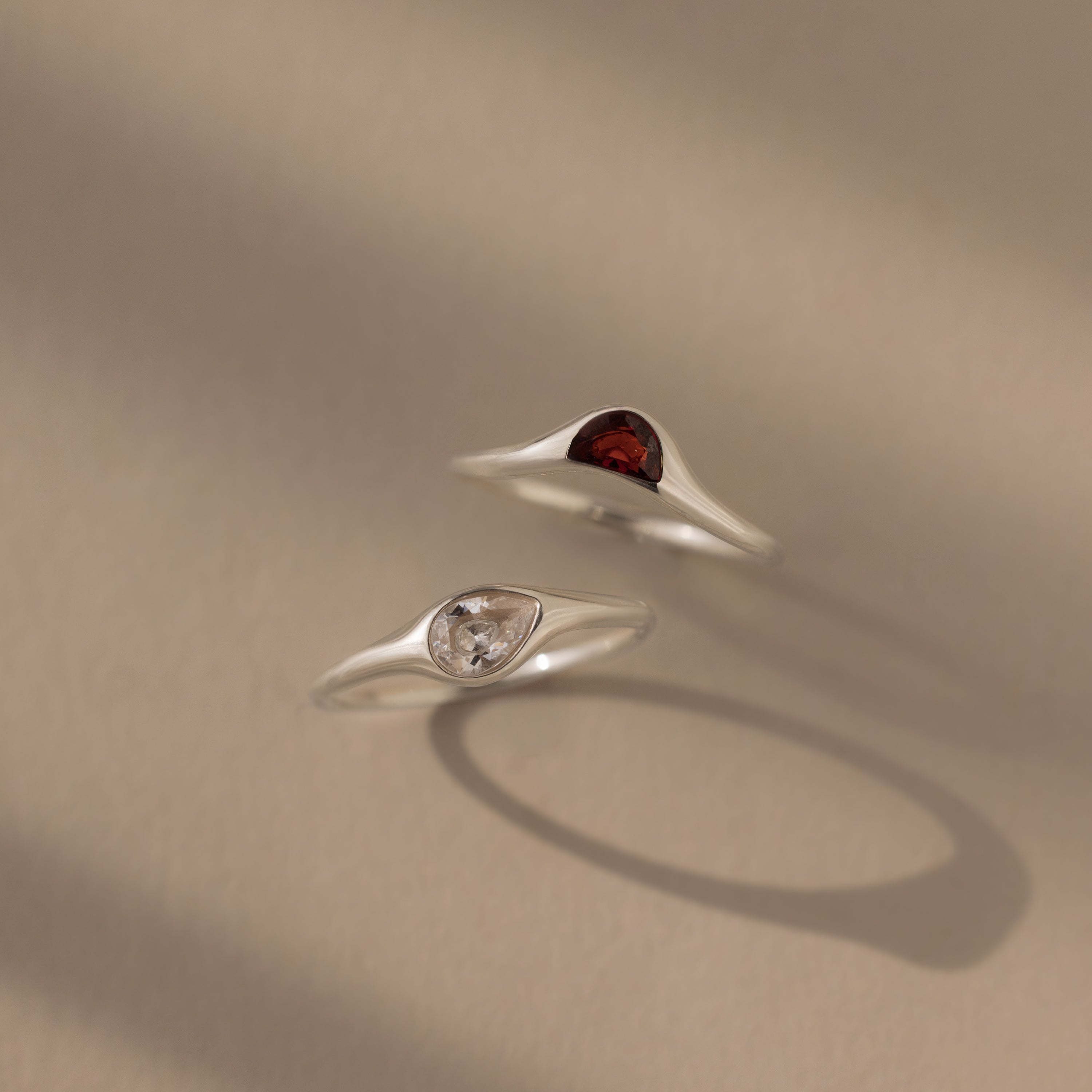 The Sculpted Toi et Moi Ring Set in Silver features two minimalist silver rings—one with a red gemstone, one with a clear stone—displayed on a neutral background.