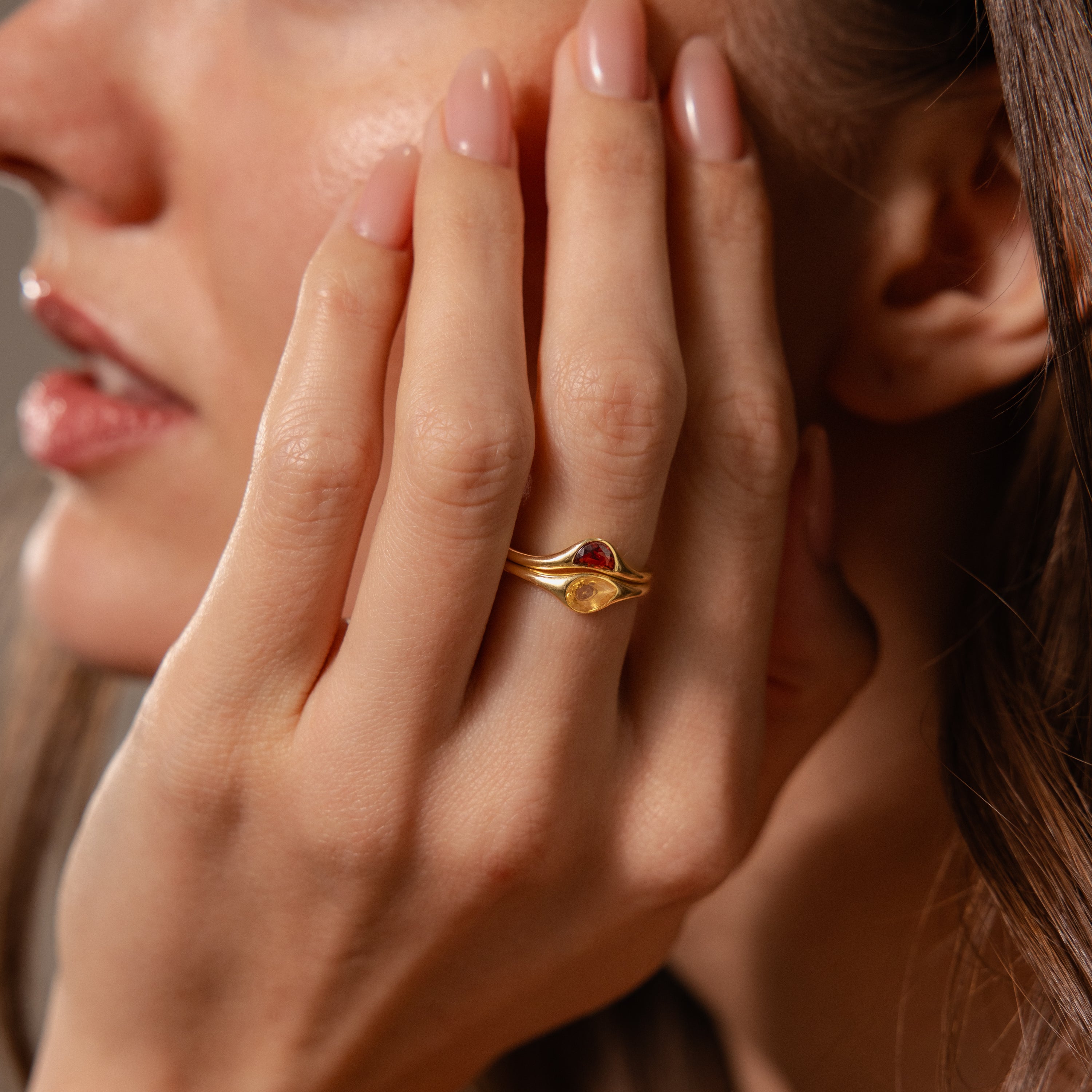A woman’s hand rests near her face, adorned with the Sculpted Toi et Moi Ring Set in 18K Gold featuring red and yellow stones. Her nails are painted with nude polish.